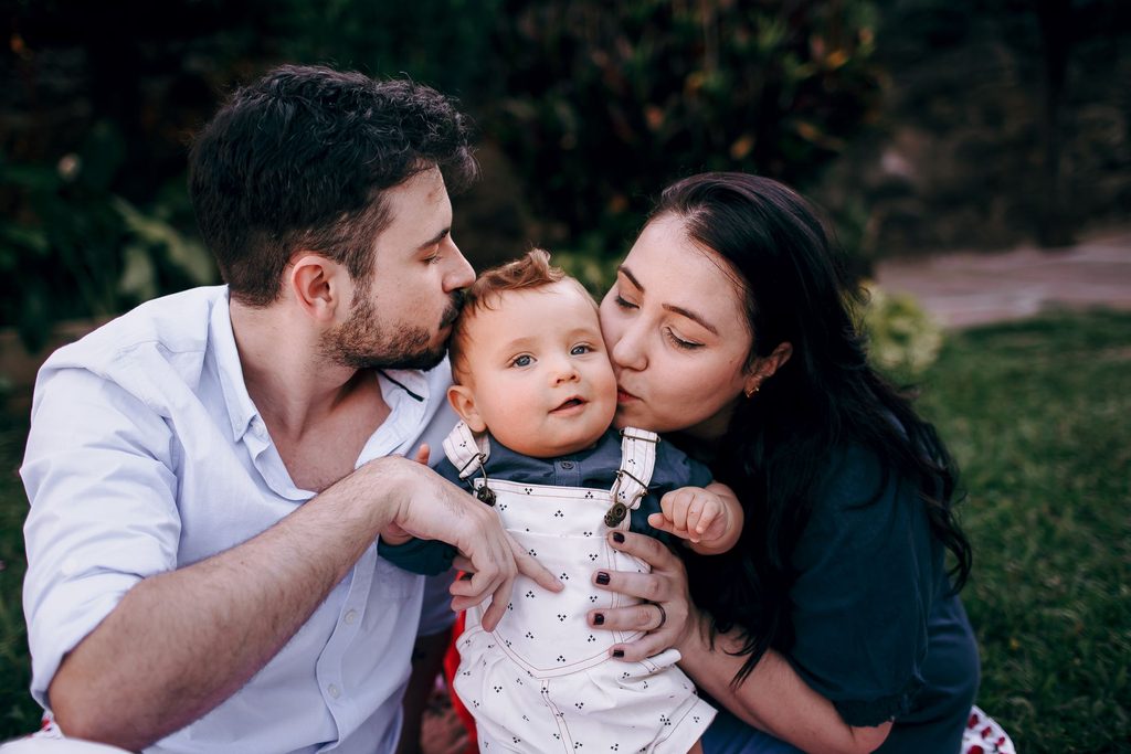 A mother and father kissing their baby on the forehead and cheek while sitting on grass