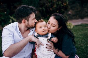 A mother and father kissing their baby on the forehead and cheek while sitting on grass